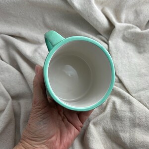 May include: A ceramic mug with a white interior and a turquoise rim and handle. The mug is held in a hand, showcasing its clean design and color contrast. The mug is empty.