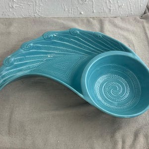 May include: A turquoise ceramic serving dish with a unique design. The dish features a curved, wing-like shape with textured details and a circular bowl. The bowl has a spiral pattern in the center. The dish is on a neutral-colored surface.