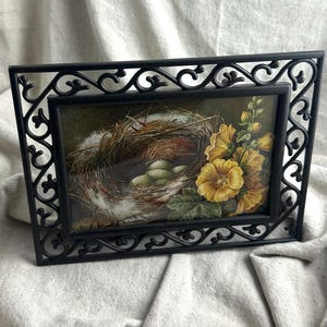 May include: A rectangular picture frame with a black metal border featuring an ornate scrollwork design. The frame contains an illustration of a bird nest with speckled eggs nestled inside, accompanied by a cluster of vibrant yellow flowers.