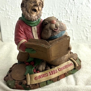May include: A Christmas figurine featuring a singing man and a beaver reading a book. The man wears a red coat, green scarf, and Santa hat. The beaver has a blue scarf. The base has a banner that reads "CARD 1995 CHRISTMAS".