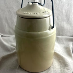 May include: A cream-colored ceramic jar with a lid and a dark blue metal handle. The lid has embossed text. The jar has a cylindrical shape with a slightly wider base. The handle arches over the top.