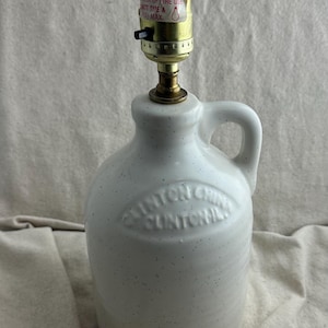 May include: A white ceramic jug lamp with a handle and a speckled finish. The lamp has a brass-colored socket and a black on/off switch. The jug has the text "CLINTON CHILE" embossed on the front.