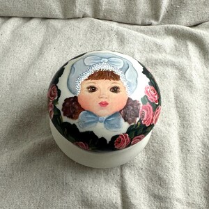 May include: A hand-painted ceramic trinket box. The lid features a portrait of a young girl wearing a blue bonnet and bow. The box is decorated with pink roses and green leaves against a black background. The base is white.