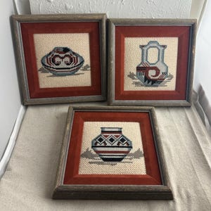 May include: Three framed needlepoint pictures of Native American pottery. The pictures are in wooden frames with red velvet mounts. The pottery is in shades of blue, white, black, and red.