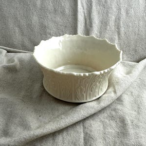 May include: Off-white ceramic bowl with a scalloped rim and a detailed leaf pattern around the base. The bowl sits on a neutral-colored fabric, showcasing its elegant design. A decorative piece for the home.