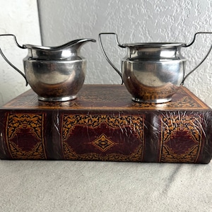 May include: A silver creamer and sugar bowl set on a brown leather-bound book with gold accents. The creamer has a handle and a spout, while the sugar bowl has two handles.