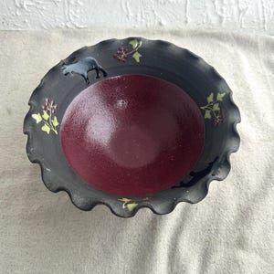 Peut inclure: Un bol en céramique gris foncé avec un bord festonné. Le bol présente un intérieur bordeaux et est décoré d'un mouton gris et d'éléments floraux jaunes et rouges. Le bol est probablement fait à la main.