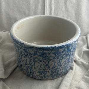 May include: A round, off-white ceramic bowl with a blue, speckled pattern. The rim and exterior are decorated with a blue design. The bowl has a simple, classic design.