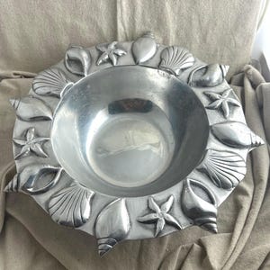 May include: A silver-colored metal bowl with a decorative border of seashells and starfish. The bowl has a circular interior and a wide, textured rim. The design evokes a coastal theme, suitable for serving or display.