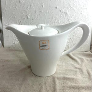 May include: A white ceramic teapot with a curved handle and spout. The teapot has a lid with a small handle and a label that reads "Luxury". The teapot is on a beige surface.