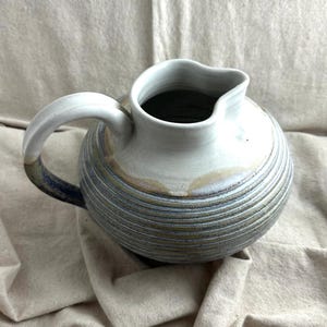 May include: A handmade ceramic pitcher with a rounded body and a curved handle. The pitcher features horizontal striped detailing in shades of blue, gray, and tan. The rim and handle are white, creating a contrast with the body.