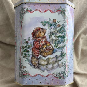 May include: A hexagonal metal tin featuring a light purple background with gold flecks. The front panel displays a watercolor illustration of a bear wearing a hat and scarf, holding a basket while standing by a stone wall in a snowy setting.