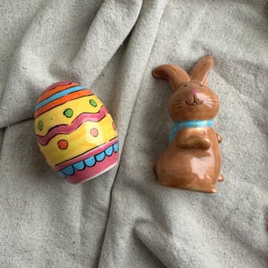May include: A brown ceramic Easter bunny wearing a blue scarf, next to a decorated Easter egg. The egg is yellow with pink, blue, orange, and green accents. The bunny has a smiling face.