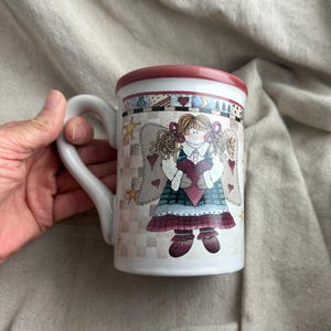 May include: White ceramic mug featuring a whimsical angel design. The mug has a red rim and handle, with a decorative band around the top. The angel illustration includes hearts, stars, and a plaid pattern, creating a charming, vintage aesthetic.