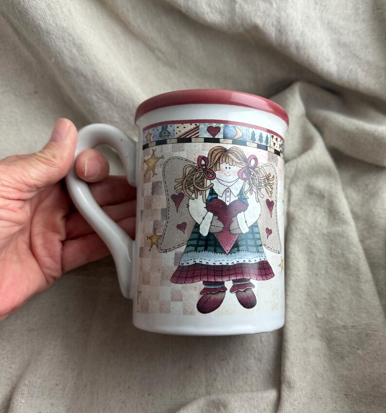 May include: White ceramic mug featuring a whimsical angel design. The mug has a red rim and handle, with a decorative band around the top. The angel illustration includes hearts, stars, and a plaid pattern, creating a charming, vintage aesthetic.