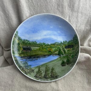 May include: A round ceramic plate featuring a hand-painted landscape scene of a blue river flowing through a green countryside with two small houses, trees, and a light blue sky with white birds. The artist signature L. Kesküla is visible.