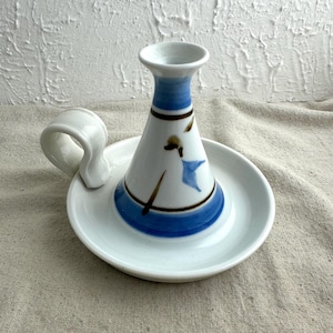 May include: A white ceramic candle holder with a blue and brown painted design. The candle holder has a circular base with a handle and a conical center. The center has a blue band and a blue and brown painted design.