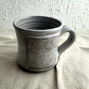 May include: Gray ceramic mug with a handle. The mug features a rectangular design with the outline of the African continent and the text "OTINO WAA EST. 2003". The mug is on a neutral background.