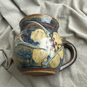 May include: A round ceramic mug featuring a mottled glaze in shades of deep blue, golden yellow, and brown. The surface displays a raised, textured botanical pattern of leaves and small berries, with a sturdy, curved handle on the right side.