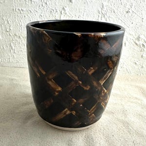 May include: A black ceramic vase with a brown criss-cross pattern. The vase has a wide mouth and a rounded bottom.