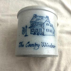 May include: A light blue stoneware jar with a lid, featuring a blue illustration of a house and the text "The Country Window." The jar has a cylindrical shape and a speckled texture, suggesting a rustic, vintage aesthetic.