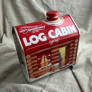 May include: A vintage Log Cabin syrup tin in the shape of a cabin, with a red and white color scheme. The tin features the text "LOG CABIN syrup" and "100th Anniversary 1887-1987". The tin has a red plastic cap.