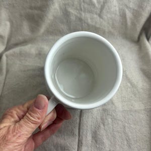 Puede incluir: Una taza de cer&aacute;mica blanca con asa, vista desde arriba. El interior de la taza tambi&eacute;n es blanco. La taza est&aacute; sostenida por una mano, con una tela de color neutro en el fondo.