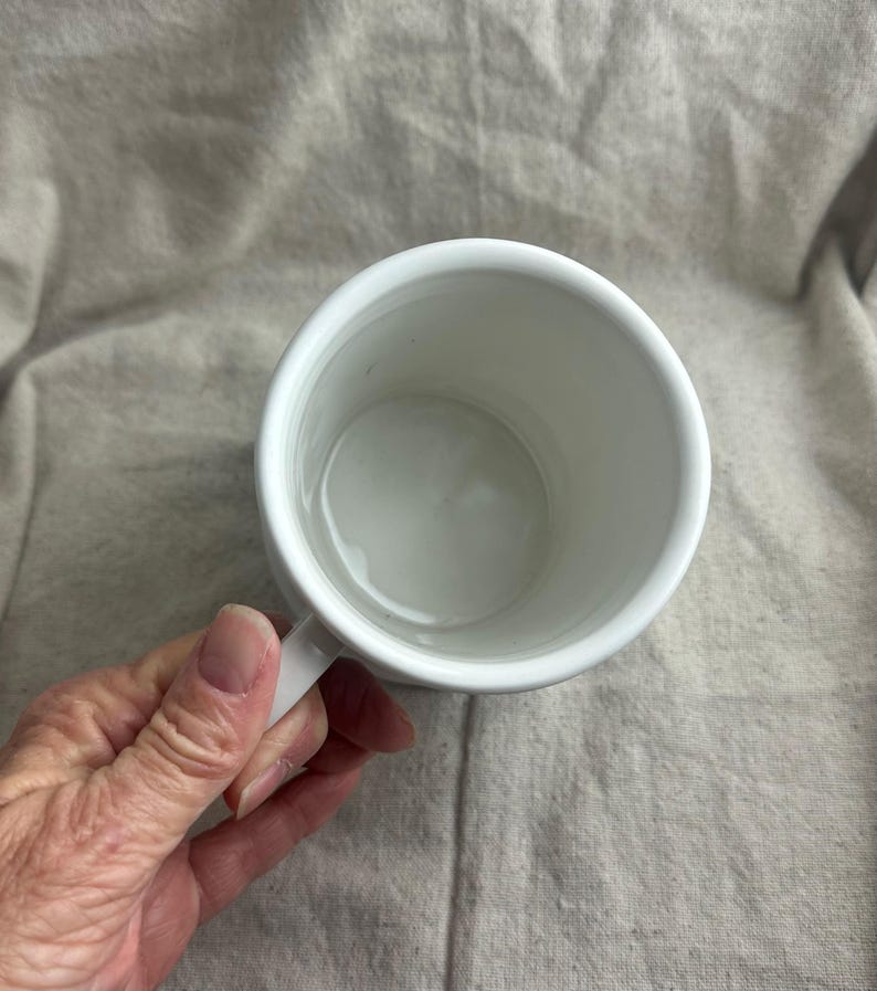 Puede incluir: Una taza de cer&aacute;mica blanca con asa, vista desde arriba. El interior de la taza tambi&eacute;n es blanco. La taza est&aacute; sostenida por una mano, con una tela de color neutro en el fondo.