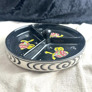 May include: A black ceramic serving dish with three compartments and a white rim. The dish is decorated with a crescent moon pattern and colorful floral designs.