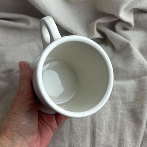 May include: A white ceramic mug with a rounded handle. The mug is held in a hand, and the interior is visible. The mug is empty and appears to be clean. The background is a neutral-colored fabric.