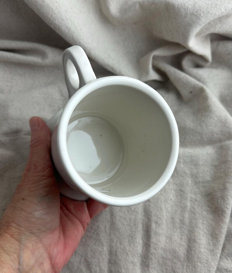May include: A white ceramic mug with a rounded handle. The mug is held in a hand, and the interior is visible. The mug is empty and appears to be clean. The background is a neutral-colored fabric.