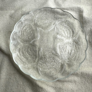 May include: Clear glass oyster serving plate with six shell-shaped compartments and a scalloped edge. The plate is designed to hold oysters and other seafood. The plate is made of clear glass.