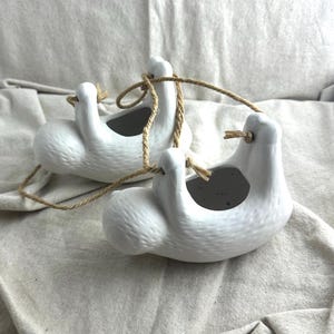 May include: Two white ceramic sloth-shaped hanging planters. Each planter has a textured surface and a circular opening for planting. They are suspended by a natural rope. The planters are designed to be decorative and functional, suitable for small plants or succulents.