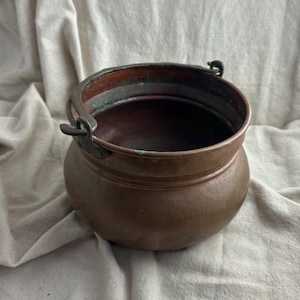May include: A round copper pot with a wide opening and a dark, weathered patina. The vessel features a sturdy, hinged metal handle attached to the sides with riveted brackets. The interior shows signs of oxidation with greenish-blue discoloration.