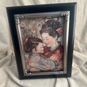 May include: A framed print featuring a mother and child in traditional Asian attire. The black frame has a silver decorative border. The print depicts the pair in a tender embrace, with floral accents in the background.