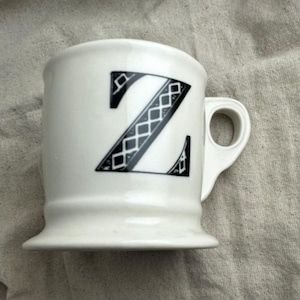 May include: White ceramic mug with a black letter "Z" design. The letter is stylized with a geometric pattern. The mug has a handle and a flared base, suitable for coffee or tea.