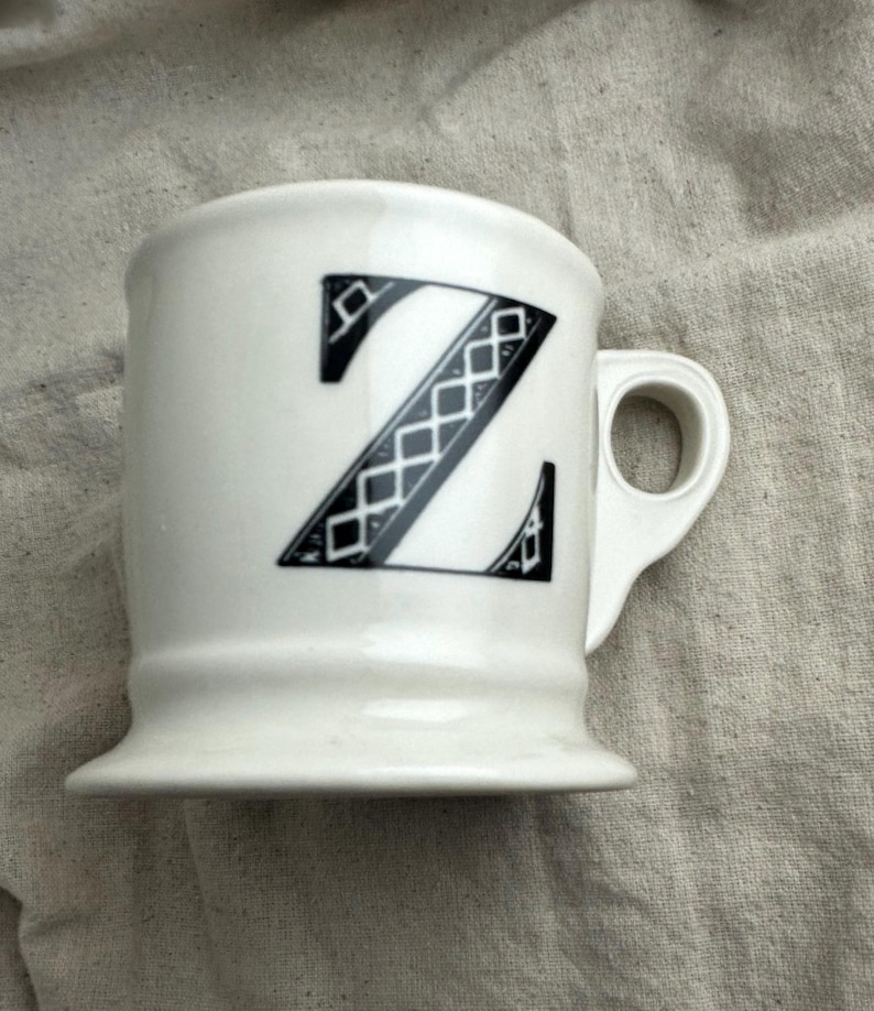 May include: White ceramic mug with a black letter "Z" design. The letter is stylized with a geometric pattern. The mug has a handle and a flared base, suitable for coffee or tea.