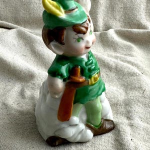 May include: A ceramic figurine of a character in a green outfit with a matching hat and a yellow feather. The figure is holding a brown object and is standing on a white cloud. The figurine has brown hair and shoes.