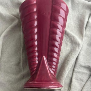 May include: A burgundy-colored ceramic vase with a unique, ribbed design. The vase has a triangular base and a flared top. The surface has a glossy finish, reflecting light. This decorative vase is suitable for floral arrangements or as a standalone art piece.