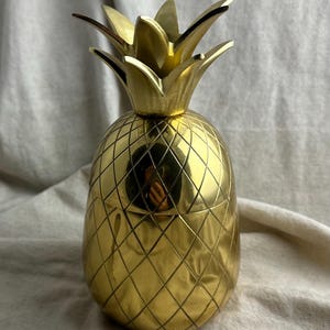 May include: A polished gold-coloured metal pineapple-shaped container with a removable lid. The body features a cross-hatched diamond pattern, and the top is adorned with pointed, stylised leaves. The surface is reflective and smooth.