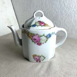 May include: White porcelain teapot with a floral design. The teapot has a lid with a small hole in the top. The floral design features pink, yellow, and blue flowers.