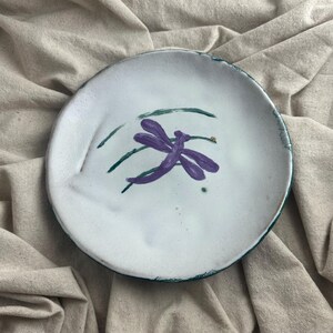 May include: A white ceramic plate with a hand-painted purple dragonfly design. The dragonfly is centered on the plate, with green lines and a small gold dot. The plate has a slightly uneven edge with a dark green trim.