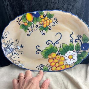 May include: An oval ceramic serving platter with a cream background and hand-painted floral design. The design features yellow and orange grapes, green leaves, white flowers, and blue accents.
