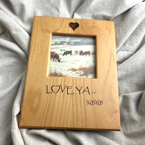 May include: A wooden picture frame with a heart-shaped cutout at the top and the words "LOVE YA... XOXO" etched into the wood. The frame holds a picture of cows in a snowy field.