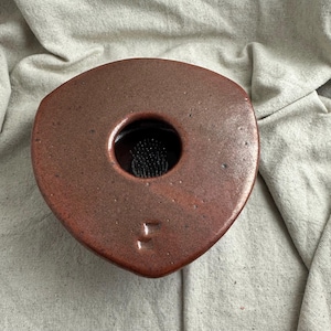 May include: A triangular, reddish-brown ceramic flower frog with a central hole. The hole contains a black floral pin holder. The frog has a smooth, slightly textured surface and is designed for arranging flowers.