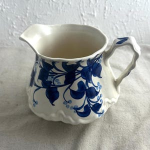 May include: A white ceramic jug with a blue floral pattern. The jug features a curved spout and a textured handle. The blue floral design adorns the body, and the base has a scalloped edge. The jug is suitable for serving liquids.