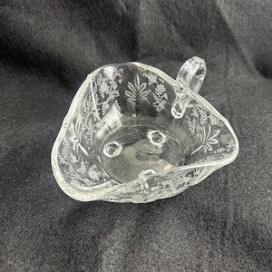 May include: Clear glass serving dish with a handle and floral etched design. The dish has three small feet and a triangular shape. The handle is arched and clear. The edges of the dish are scalloped.