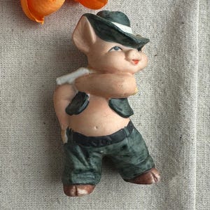 May include: A ceramic figurine of a pig wearing a green and black cowboy hat and vest, and green pants. The pig is standing with its arms crossed and has a friendly expression.