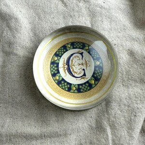 May include: A clear glass paperweight with a decorative design. The design features a white background with a blue and gold letter "C" in the center, surrounded by a green and gold pattern.