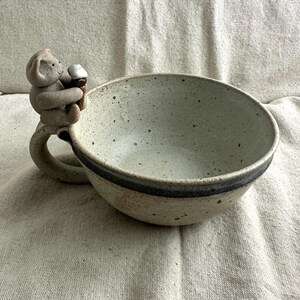 May include: A speckled ceramic bowl with a monkey-shaped handle. The monkey is holding an ice cream cone. The bowl has a dark gray band around the rim and is light beige with dark speckles.